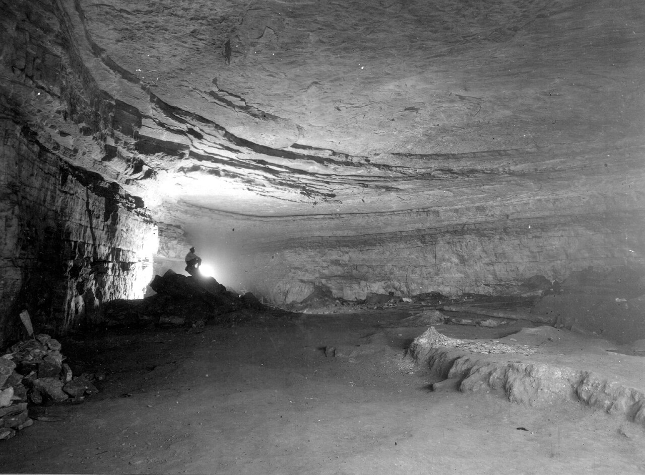 A black and white historical photograph showing a large, wide cavern inside Mammoth Cave National Park with a person standing near the entrance.