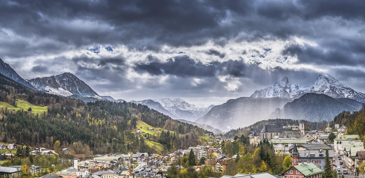 A panoramic view of a town nestled in a valley beneath dramatic, dark, storm-like clouds over mountains.