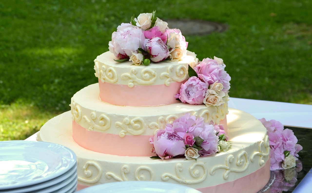 Three-tier white wedding cake with cream scroll piping and pink peony and rose decorations, outdoors on a table.