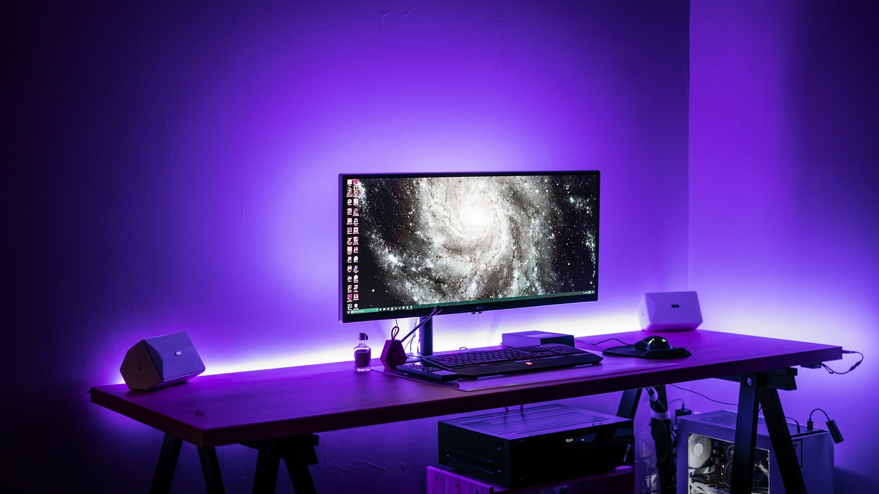A gaming desk setup with a computer monitor displaying a galaxy image, illuminated by purple LED lights.