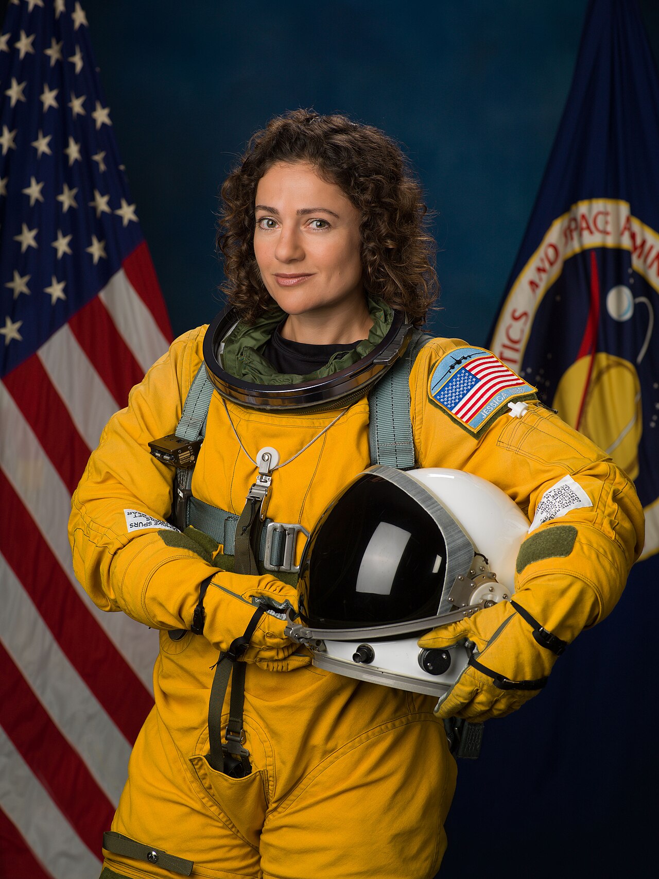 Portrait of astronaut Jessica Meir in a yellow flight suit holding a helmet, with US and NASA flags in the background