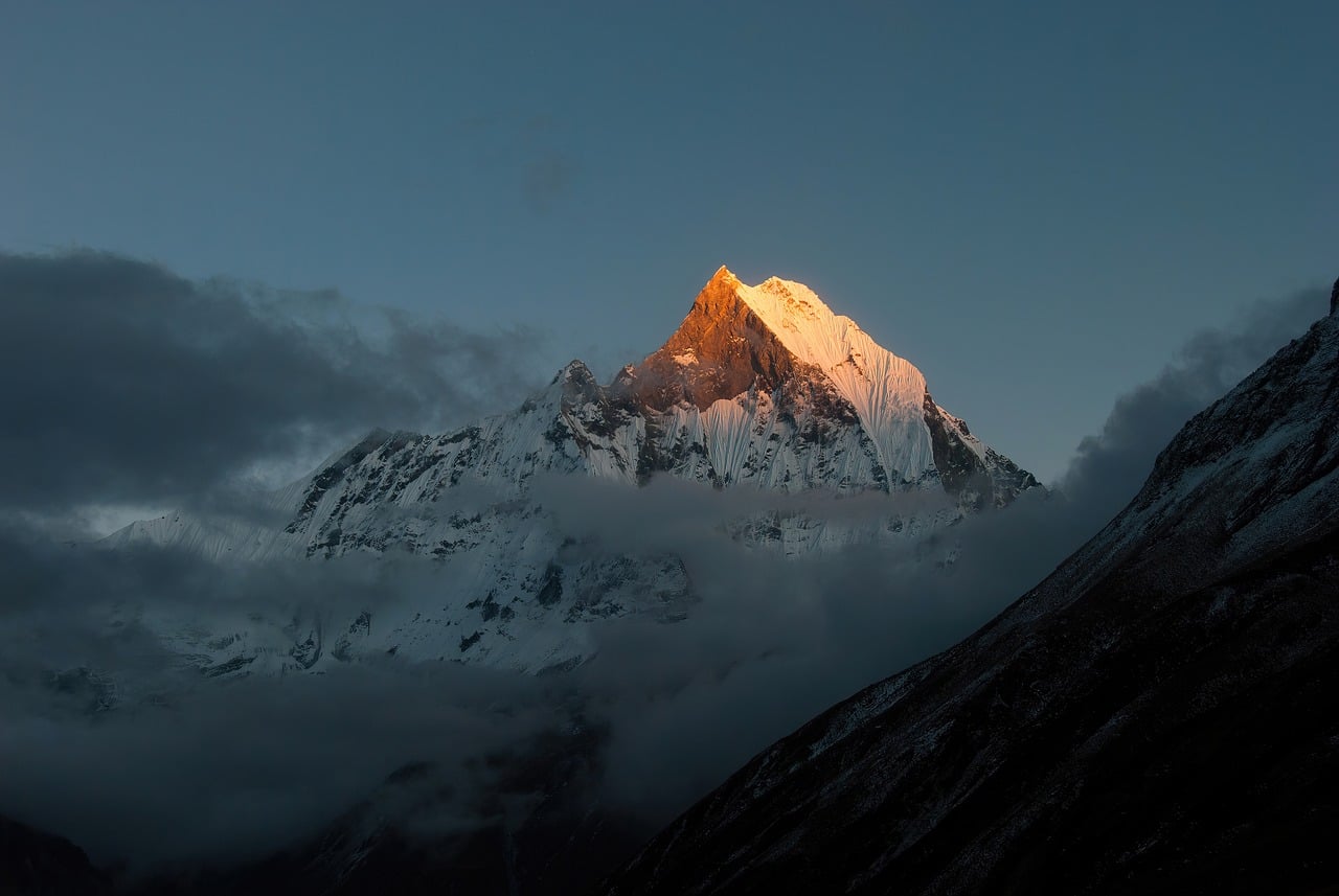 A majestic, snow-capped mountain peak illuminated by the golden light of sunrise against a dark blue sky.