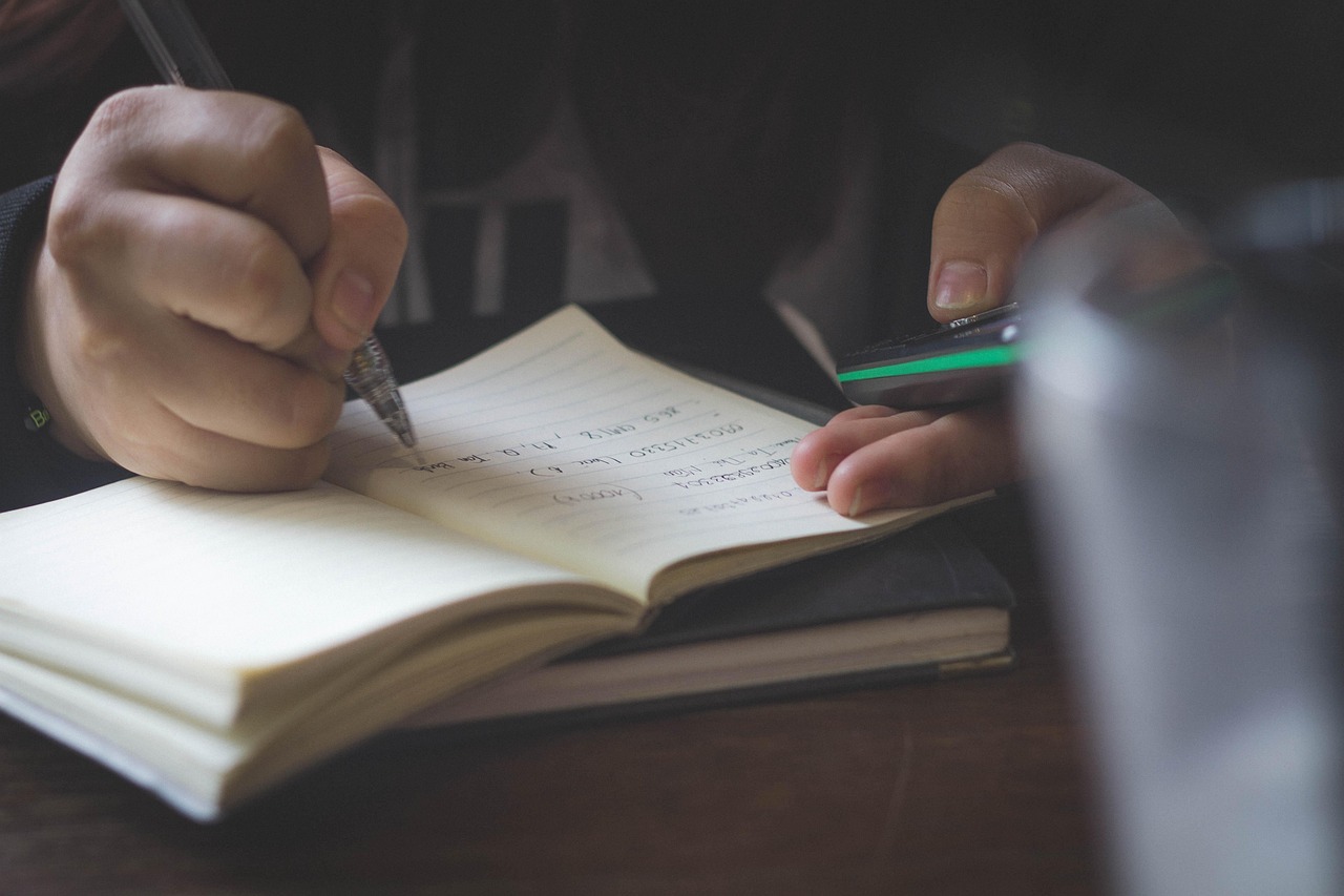 A person writing notes in a notebook with a pen while holding a calculator