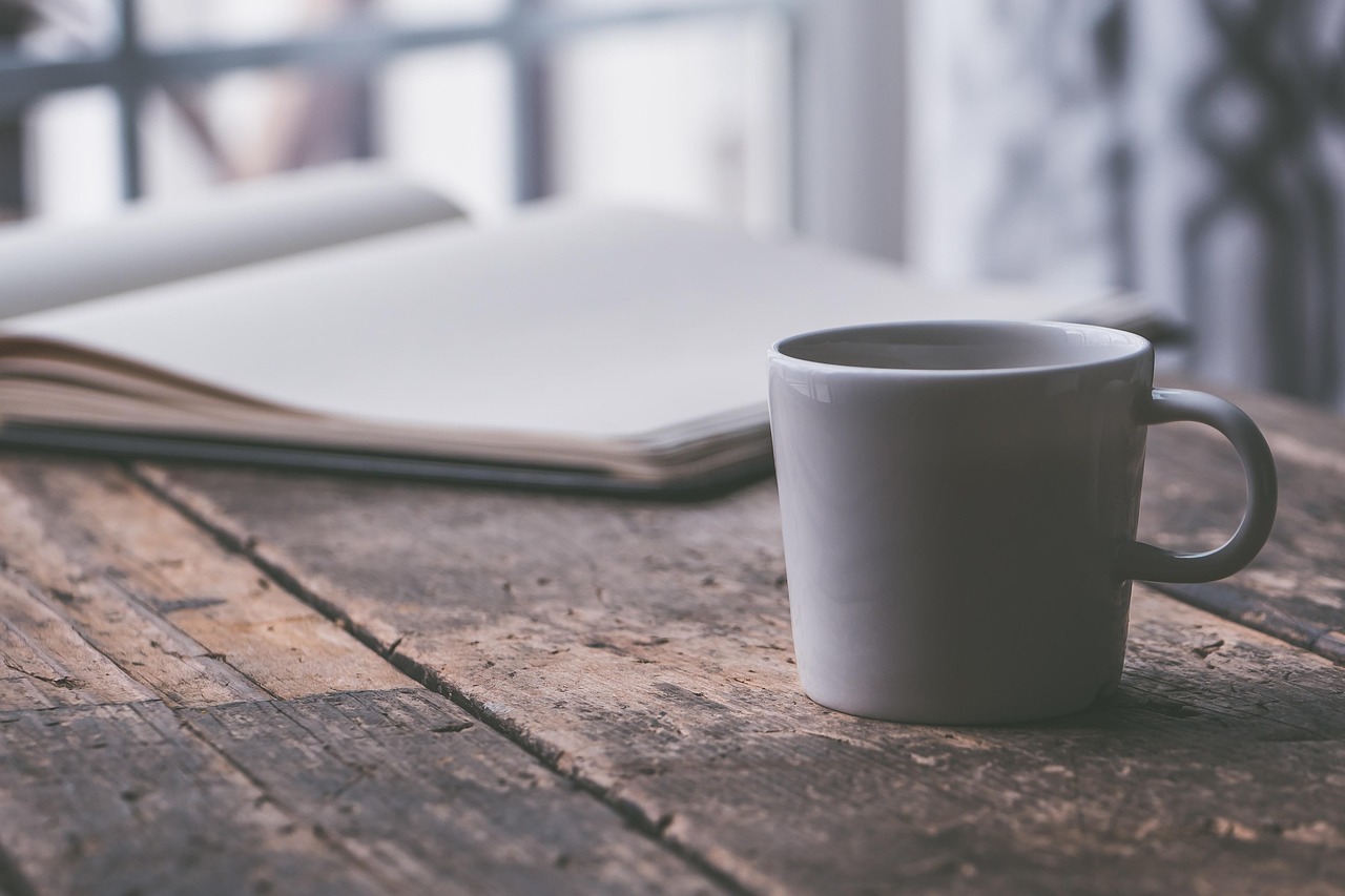 A white ceramic coffee cup on a rustic wooden table with an open notebook in the background.