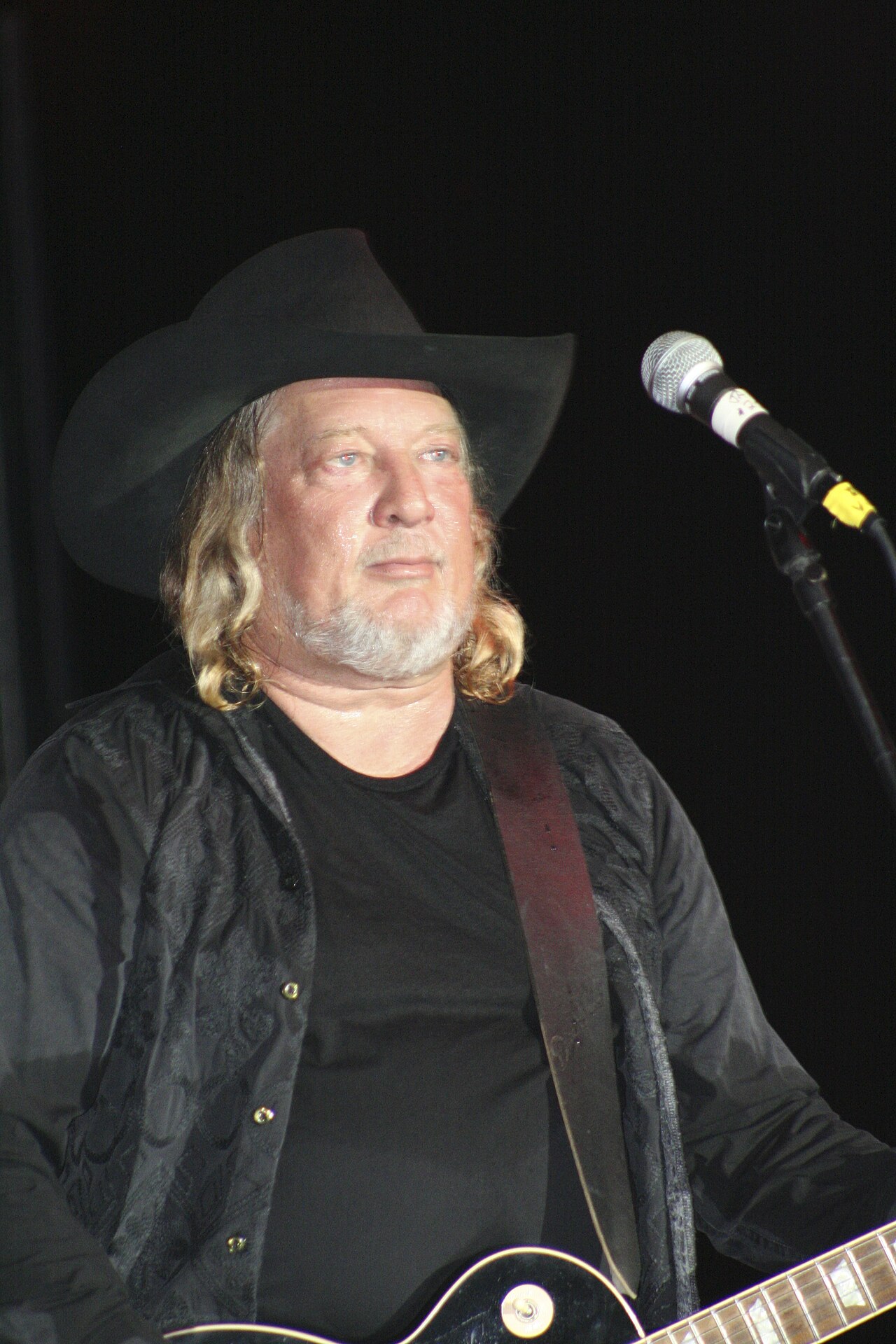 John Anderson performing on stage in a black cowboy hat, with a guitar