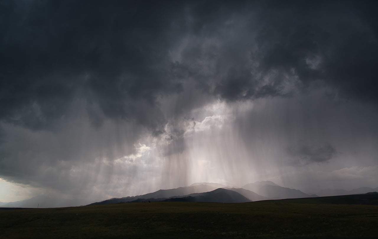 A dark, stormy sky with heavy rain falling over a landscape of hills.