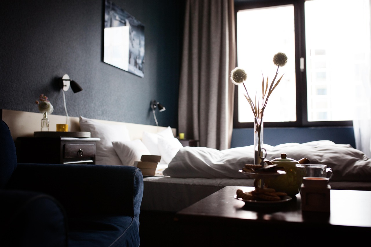 A cozy, well-lit hotel bedroom with a comfortable bed and a small table with snacks and a vase of flowers.