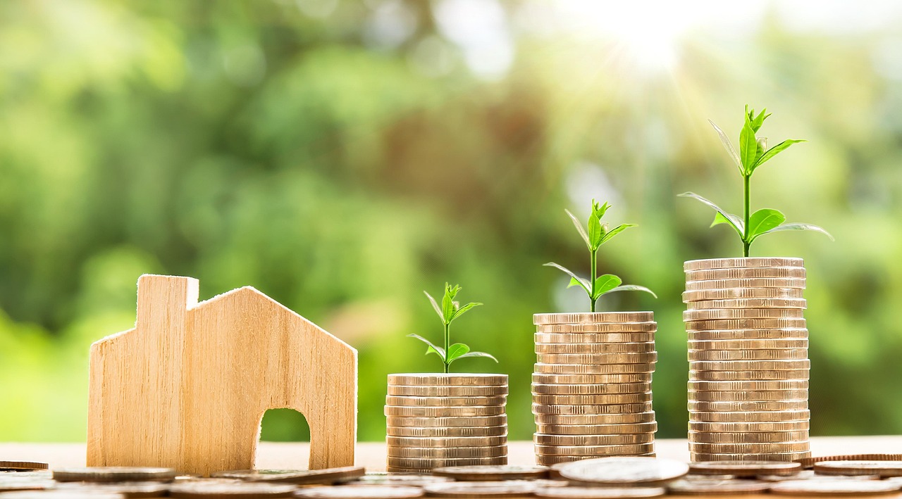 A small wooden house model next to stacks of coins with small green plants growing out of them, symbolizing financial growth and real estate investment.