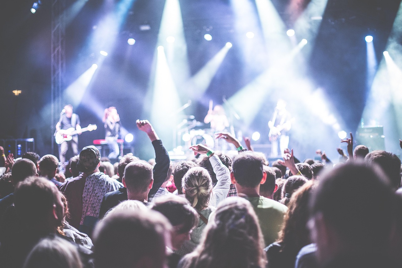 A crowd of people at a concert watching a band perform on a brightly lit stage.