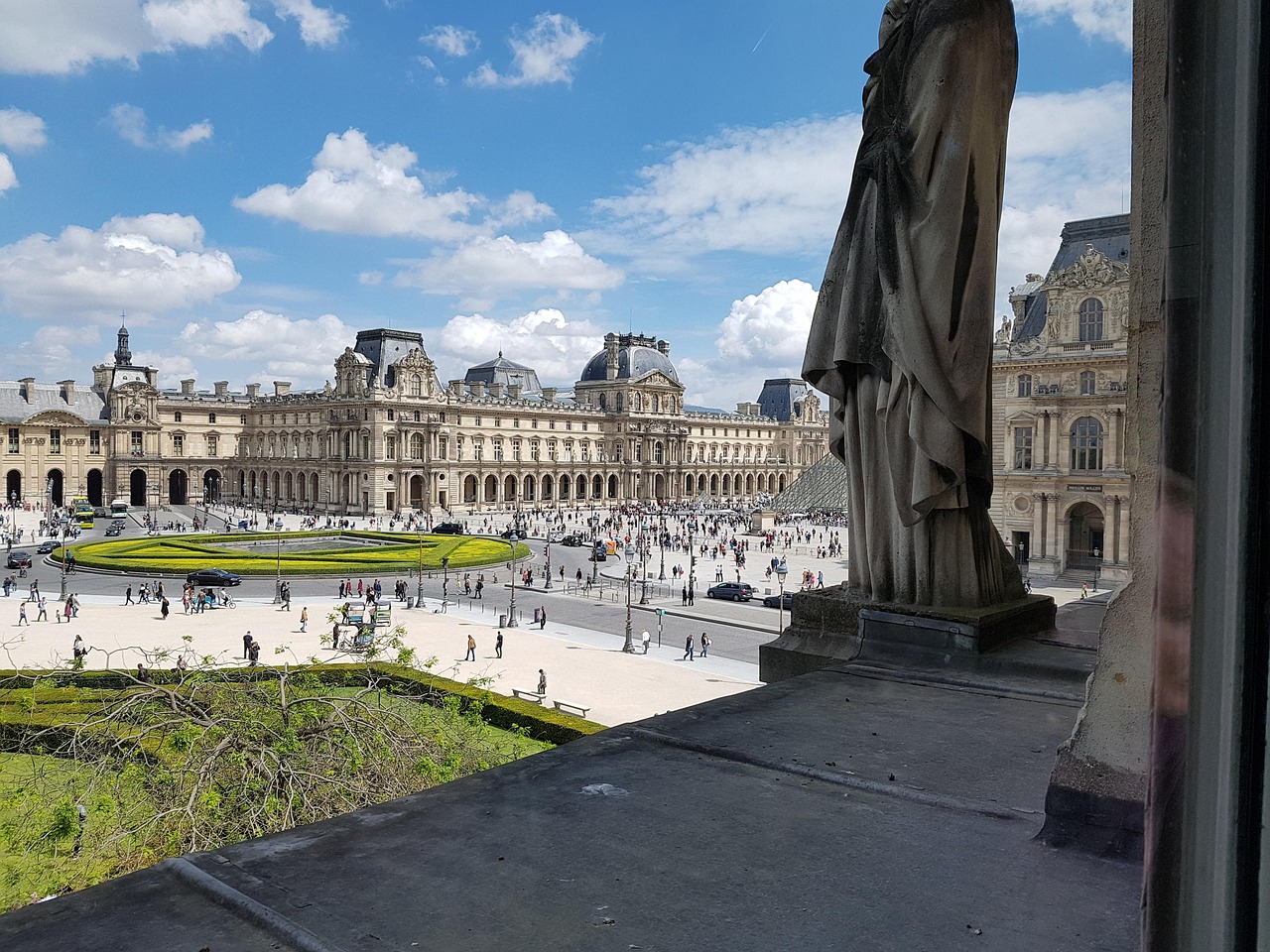 A scenic view of a historic Parisian square and architecture resembling the Louvre museum, viewed from an elevated window with a stone statue in the foreground.