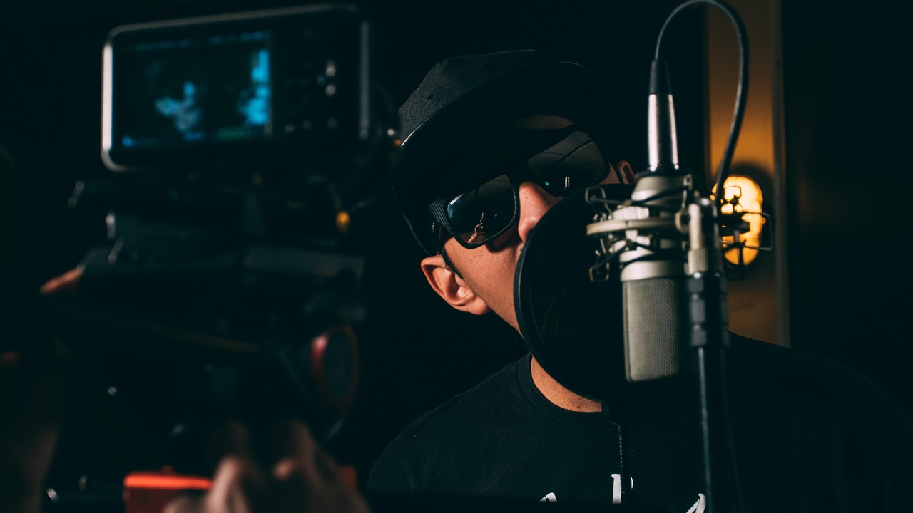 A musician singing into a studio microphone in front of a camera.