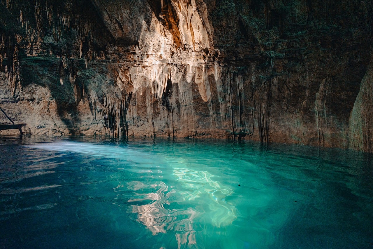 A serene underground pool of clear turquoise water inside a cave with stalactites hanging from the ceiling.