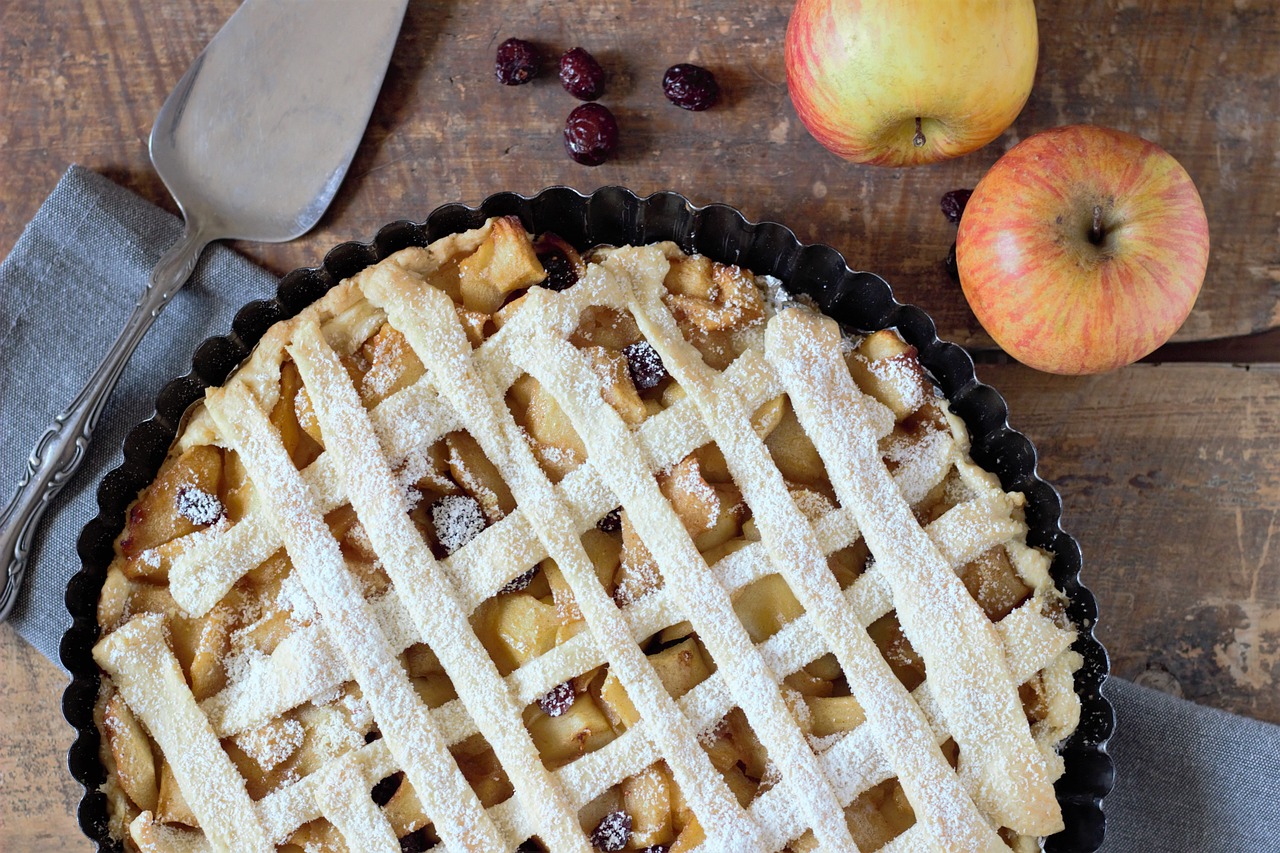 A freshly baked apple tart with a lattice crust, dusted with powdered sugar, sitting on a wooden table next to two whole apples.