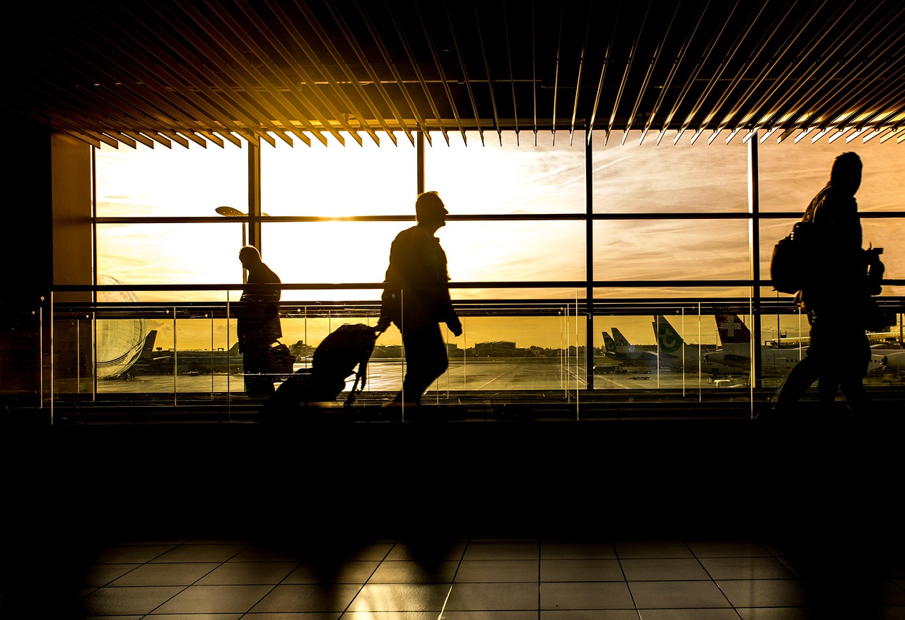 Silhouettes of travelers walking through an airport terminal with airplanes visible through the windows at sunset.