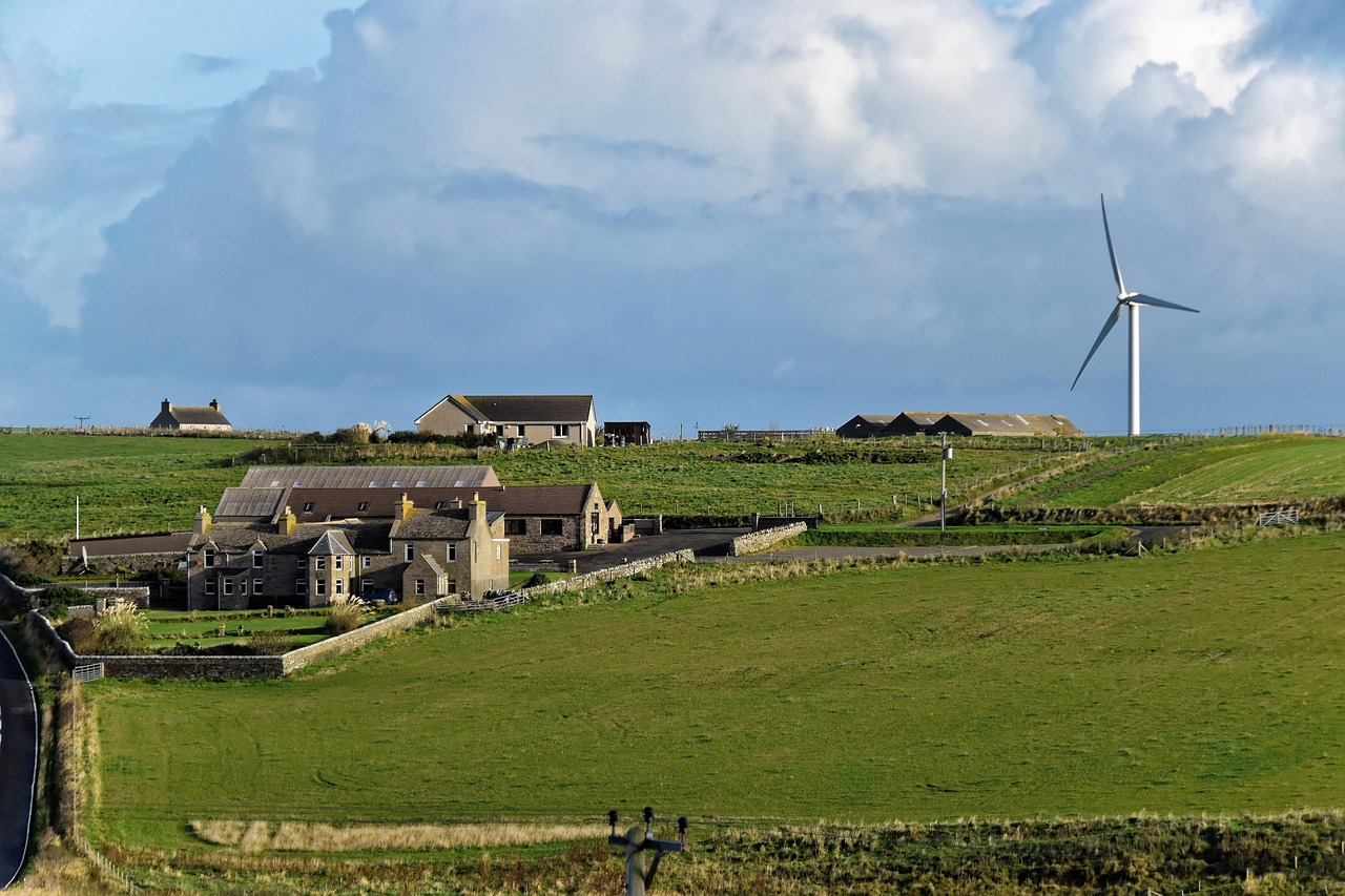 A rural landscape in the UK featuring green fields, farm buildings, and a wind turbine under a cloudy sky.