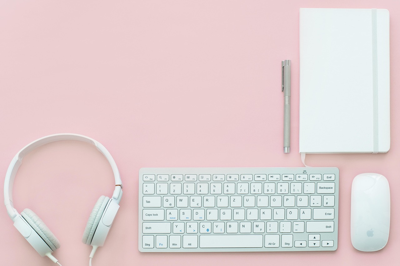 A pair of white headphones, a computer keyboard, a mouse, a pen, and a notebook arranged on a pink background.