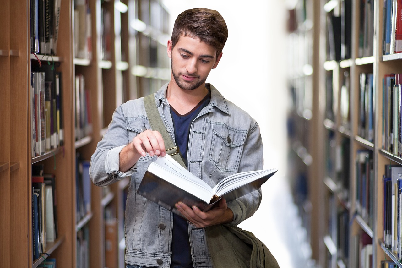 A young male student standing in a library aisle, reading a book.