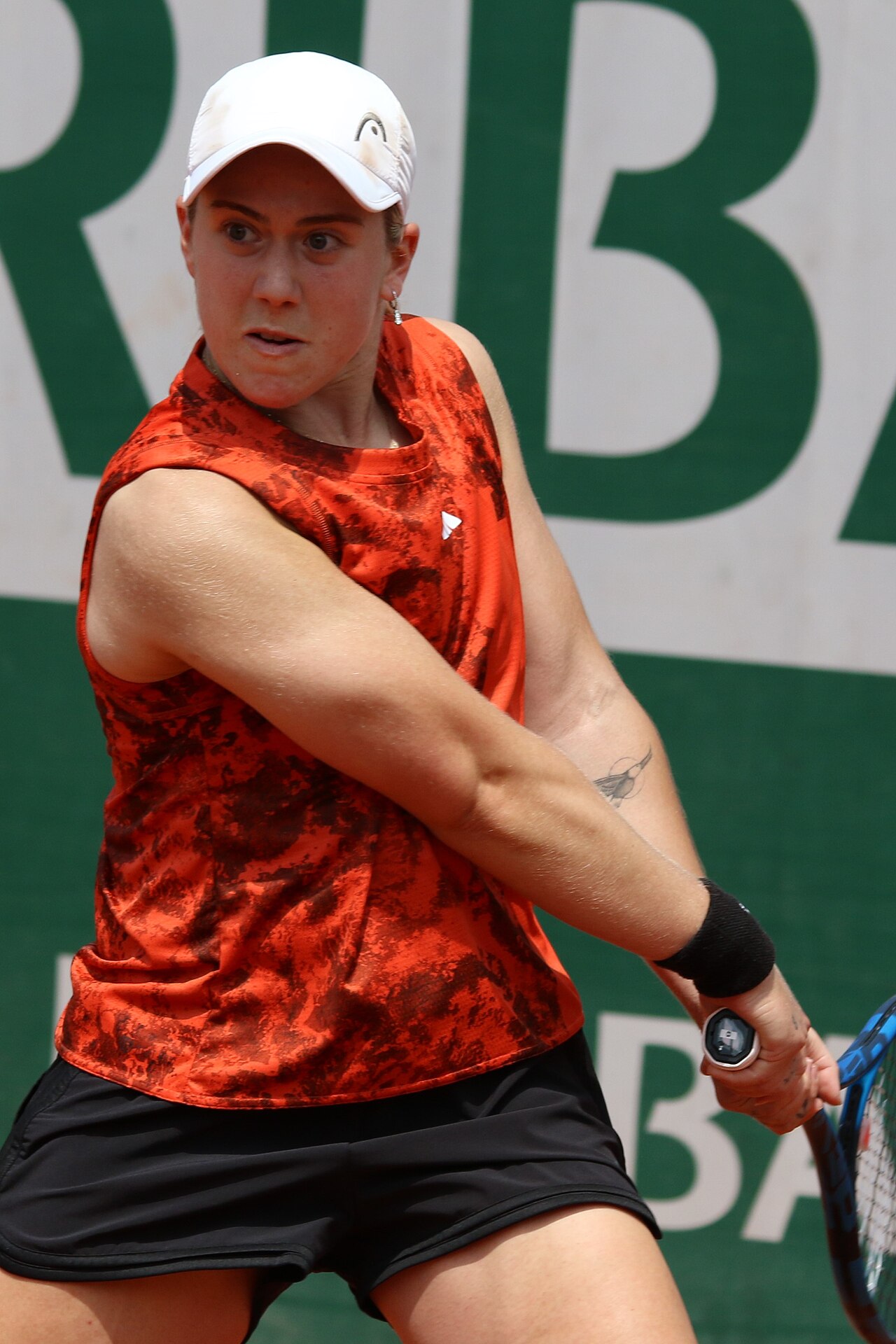 Sonay Kartal playing tennis in an orange top and white cap.